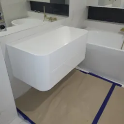 Biała, podwieszana szafka łazienkowa z frezowanymi frontami, złota armatura, marmurowe płytki i zabezpieczona podłoga podczas montażu.