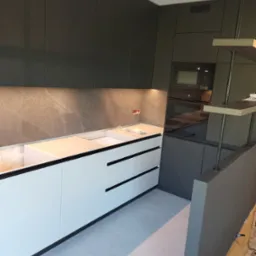 Nowoczesna kuchnia z białymi dolnymi szafkami i ciemnoszarymi górnymi, z marmurowym blatem i wbudowanym piekarnikiem, oświetlona listwą LED pod szafkami.