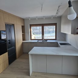 NomadMeble Adam Tatar - Nowoczesna kuchnia z wyspą, fronty w odcieniach beżu i drewna, marmurowy blat, czarna lodówka, oświetlenie szynowe. Widok na zimowy krajobraz za oknem.