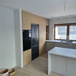 NomadMeble Adam Tatar - Nowoczesna kuchnia z drewnianymi szafkami, czarną lodówką i piekarnikiem w zabudowie. Wyspa kuchenna z białym blatem. Okno z widokiem na zewnątrz. Podłoga drewniana.