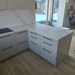 NomadMeble Adam Tatar - Jasnoszara kuchnia na wymiar z blatem imitującym marmur i czarnymi uchwytami. W tle okno z widokiem na zimowy krajobraz i mężczyzna z telefonem.