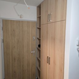 NomadMeble Adam Tatar - Jasna szafa wnękowa z półkami obok drzwi wejściowych, dopasowana kolorystycznie do drzwi. Minimalistyczny design, widoczne metalowe uchwyty.