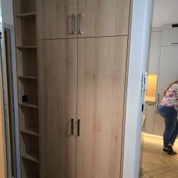 NomadMeble Adam Tatar - Jasna, drewnopodobna szafa z półkami i drzwiami, dopasowana do wnętrza z parkietem w jodełkę. Minimalistyczne uchwyty i oświetlenie LED w tle.