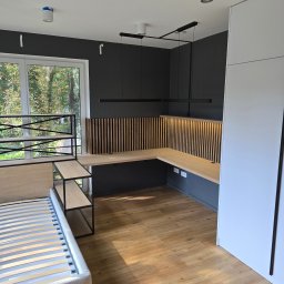 NomadMeble Adam Tatar - Nowoczesny pokój z biurkiem w kształcie litery L, drewnianą ścianką działową z lamelami i łóżkiem z metalowym regałem. Minimalistyczne oświetlenie sufitowe.
