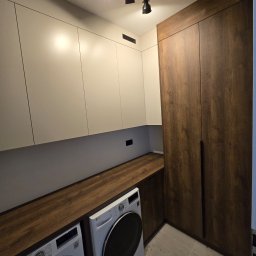 NomadMeble Adam Tatar - Nowoczesna aranżacja pralni z zabudową meblową: białe szafki wiszące, drewniany blat nad pralkami i wysoka szafa w odcieniu ciemnego drewna. Oświetlenie punktowe.