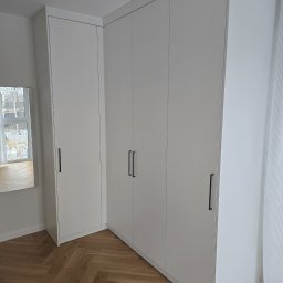 Biała, narożna szafa wnękowa z minimalistycznymi, czarnymi uchwytami, na parkiecie ułożonym w jodełkę. W tle widoczne okno i lustro.