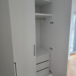 Biała szafa z otwartymi drzwiami, widoczne półki, drążek na ubrania i szuflady. Minimalistyczny design, jasne wnętrze, parkiet na podłodze.