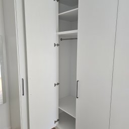Biała szafa wnękowa z otwartymi drzwiami, widoczne półki i drążek na ubrania. Minimalistyczny design, czarne uchwyty, parkiet na podłodze.