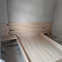 Drewniane łóżko z szafkami nocnymi w surowym wnętrzu. Widoczne gniazdka elektryczne i fragmenty folii ochronnej. Jasne drewno, minimalistyczny design.