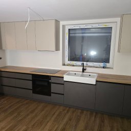 NomadMeble Adam Tatar - Nowoczesna kuchnia: szare szafki dolne z drewnianym blatem, biała umywalka farmerska, beżowe szafki górne. Widoczne gniazdka elektryczne i niedokończone wykończenie.