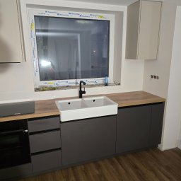 NomadMeble Adam Tatar - Szara zabudowa kuchenna z drewnianym blatem i białym zlewem farmerskim pod oknem z ramą Kömmerling. Widoczne szafki wiszące i sprzęt AGD.