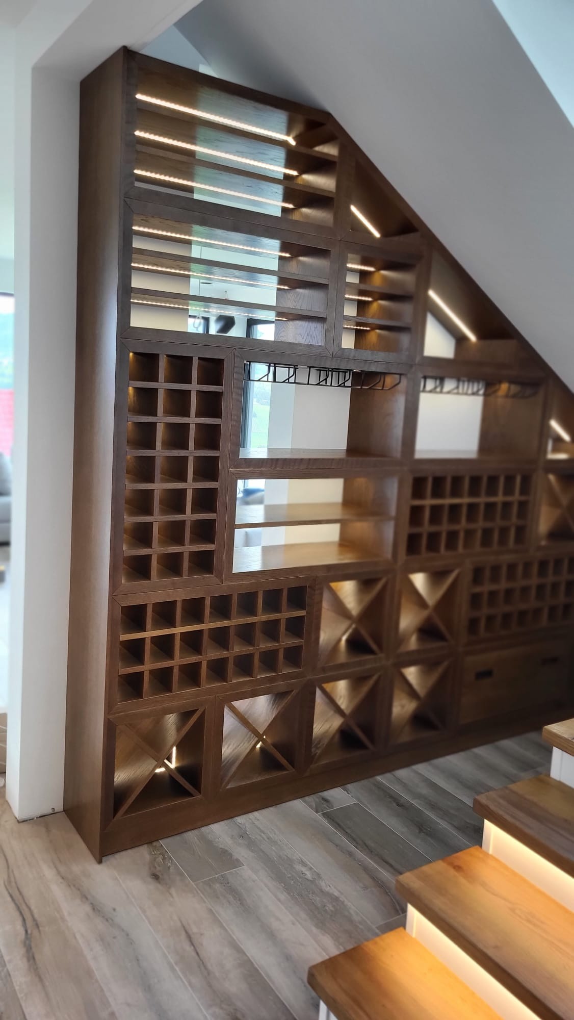Drewniana zabudowa pod schodami z podświetleniem LED, tworząca regał z przegródkami na wino i szklanki. Widoczne schody z oświetleniem.