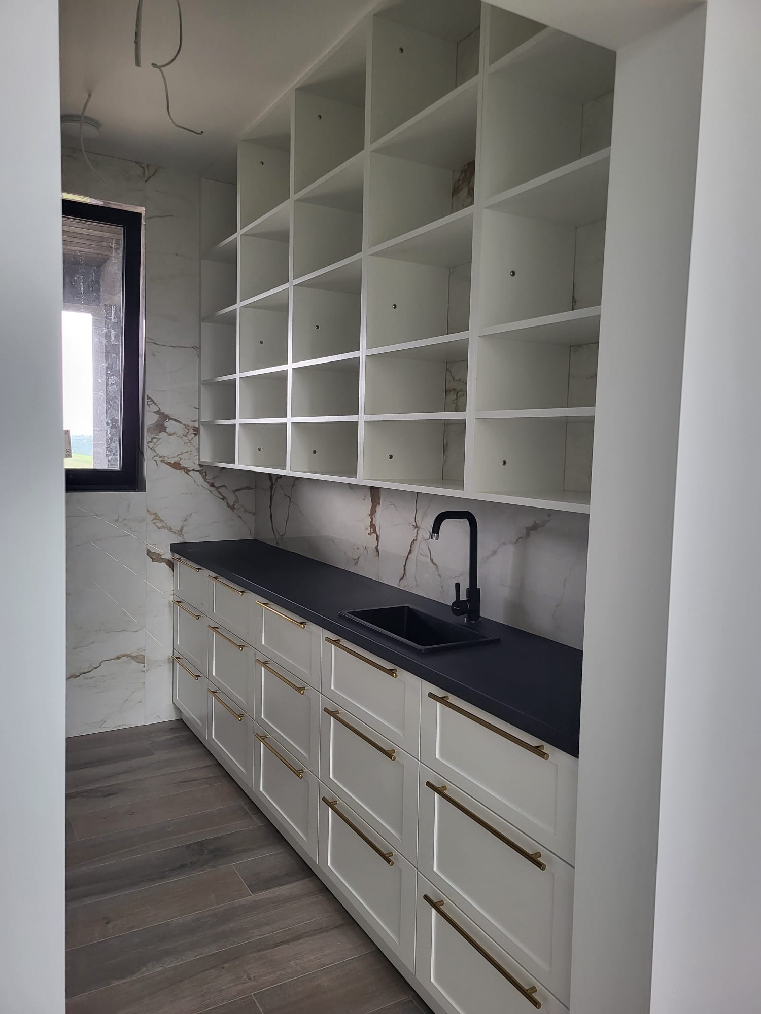 Białe szafki kuchenne z czarnym blatem i złotymi uchwytami, uzupełnione o regał z kwadratowymi półkami. Marmurowe płytki na ścianie, drewnopodobna podłoga.