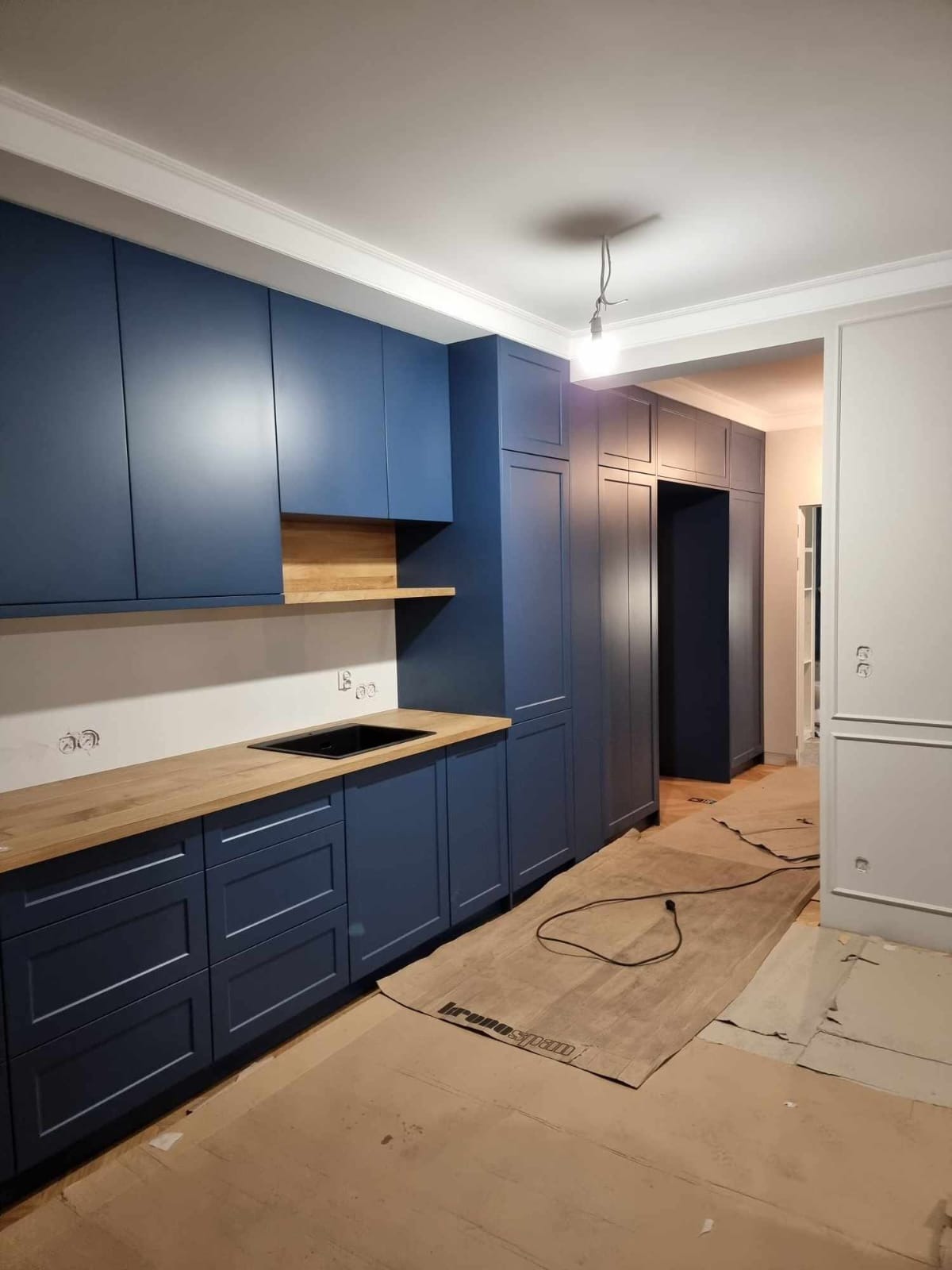 Granatowa zabudowa kuchenna z drewnianym blatem i półkami w Wadowicach. Widoczna również szafa wnękowa w korytarzu. Oświetlenie sufitowe w trakcie montażu.