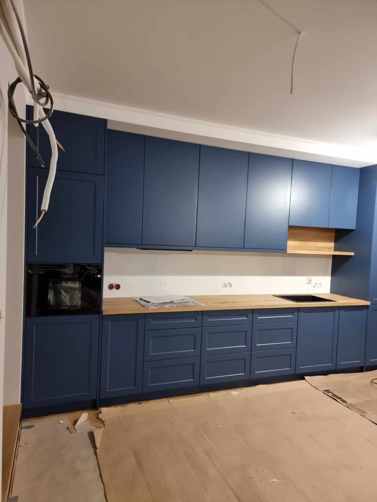 Granatowa zabudowa kuchenna z drewnianym blatem, widoczny piekarnik i płyta grzewcza. Częściowo zakryta podłoga. Wiszące kable. Nowoczesny styl.