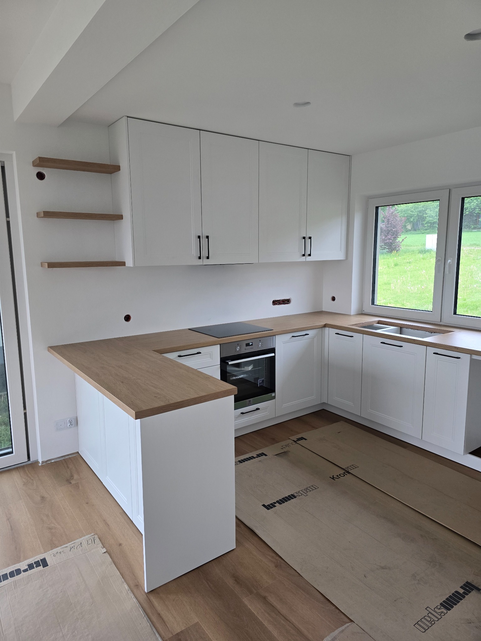 Jasna kuchnia z drewnianym blatem i białymi szafkami, minimalistyczny design, widok na ogród przez okno, osłonięta podłoga, półki na ścianie.