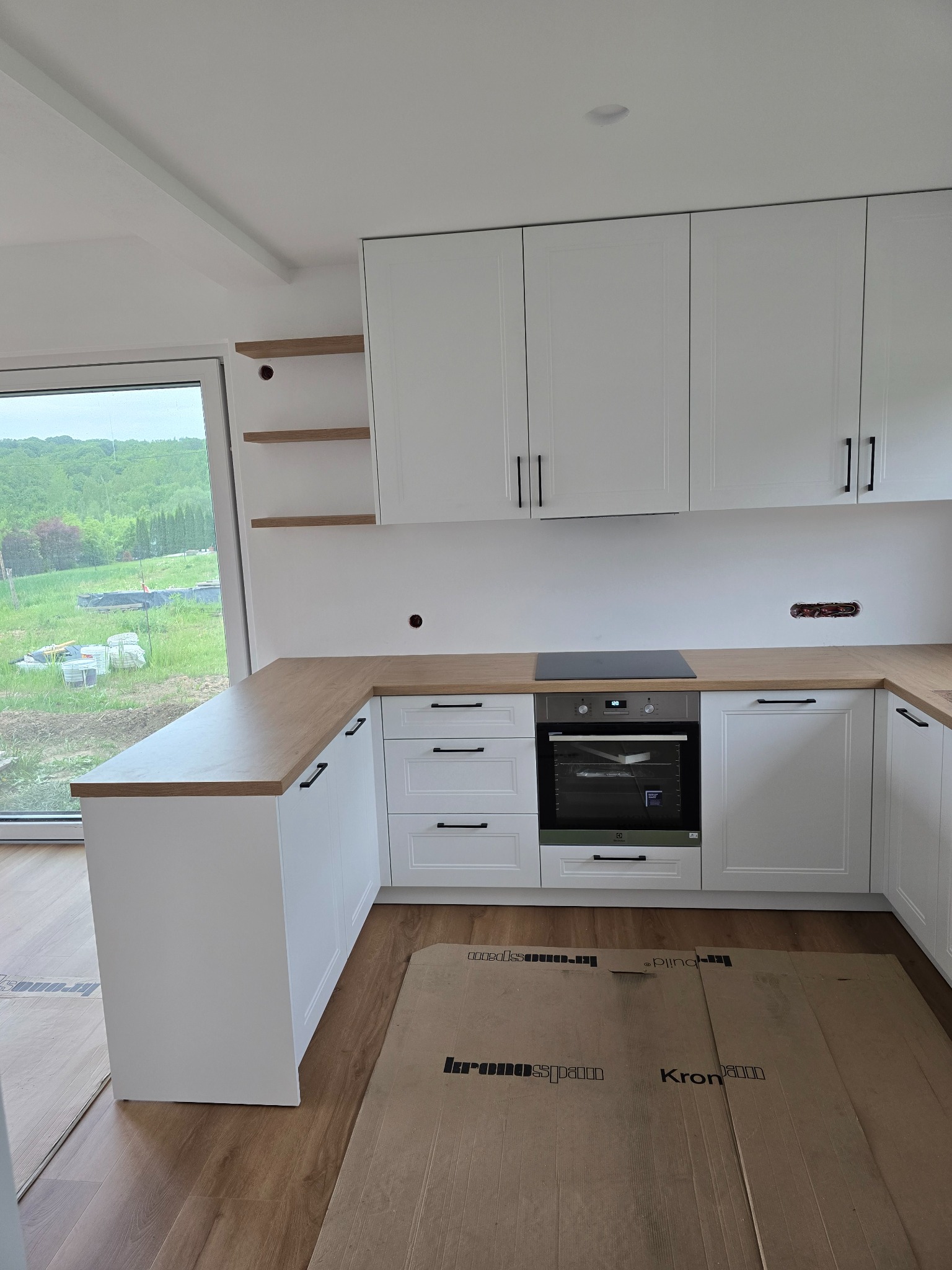 Jasna kuchnia z drewnianym blatem, minimalistyczne fronty, czarne uchwyty. Widoczne niedokończone wykończenia i zabezpieczenie podłogi kartonem.