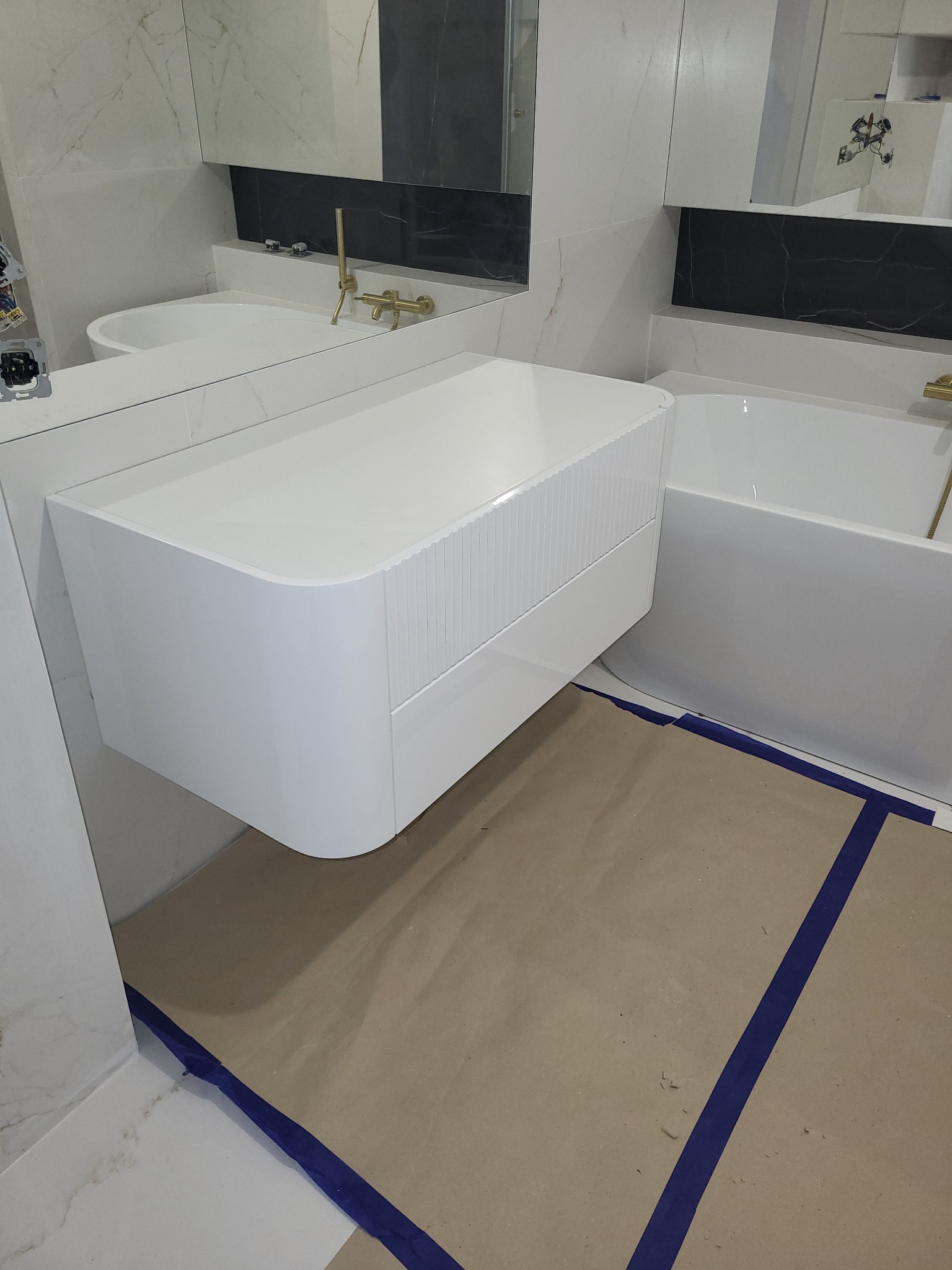 Biała, podwieszana szafka łazienkowa z frezowanymi frontami, złota armatura, marmurowe płytki i zabezpieczona podłoga podczas montażu.