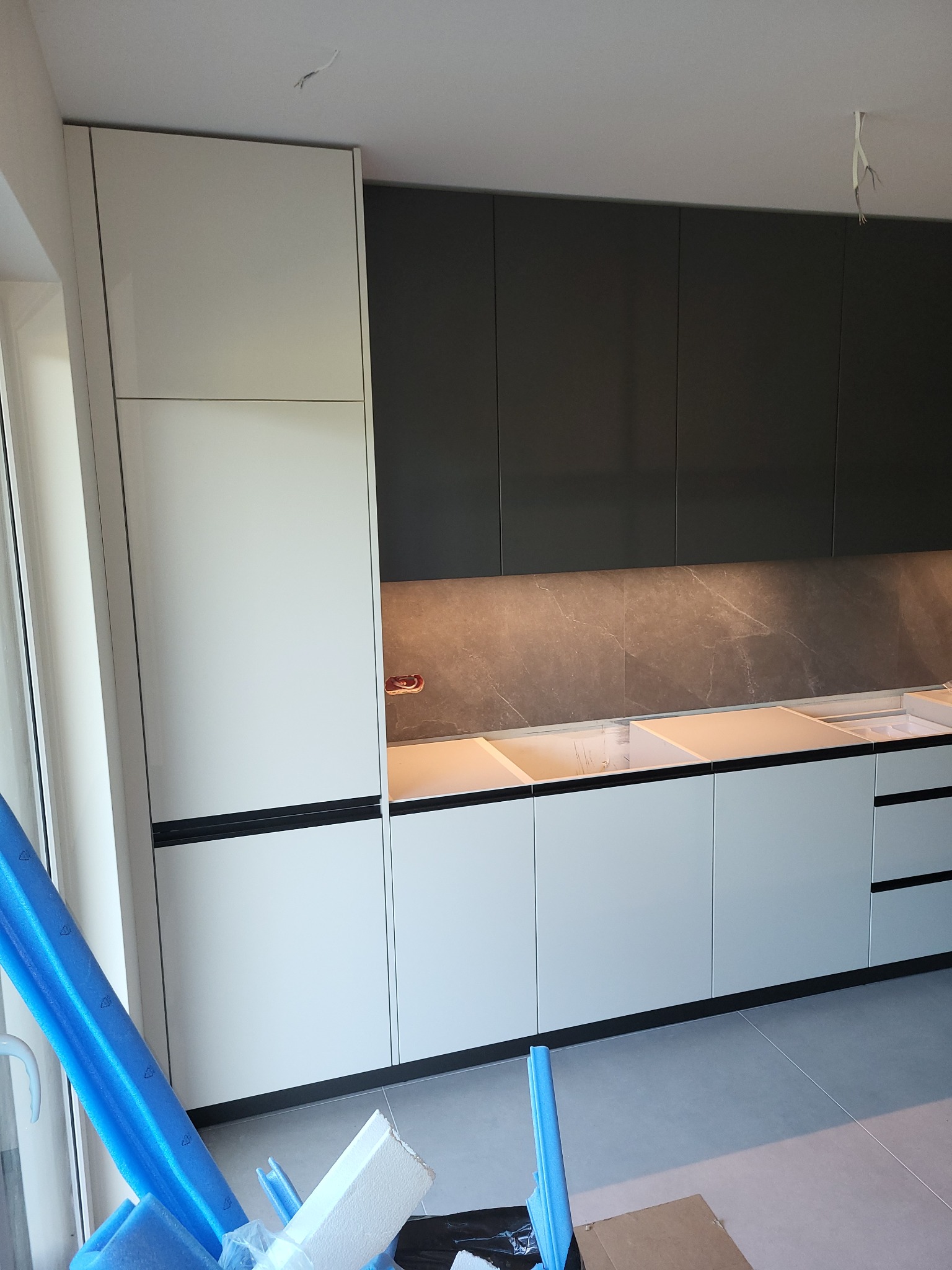 Nowoczesna kuchnia z białymi i grafitowymi szafkami, marmurowy backsplash i oświetlenie LED, częściowo zasłonięta materiałami zabezpieczającymi podczas montażu.