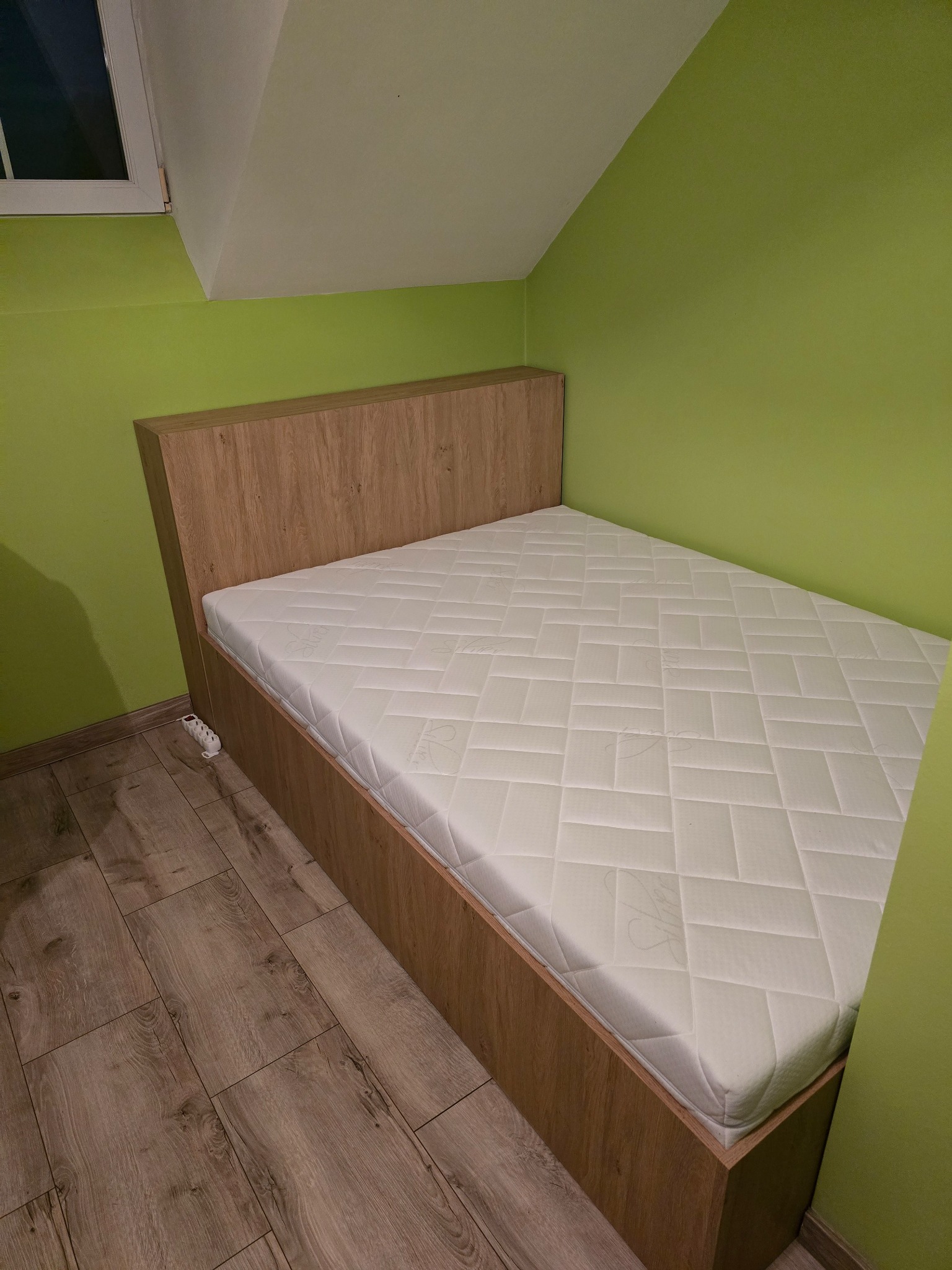 Drewniane łóżko z materacem w pokoju z zielonymi ścianami i skośnym sufitem. Widoczna podłoga z paneli. Minimalistyczny design, jasne wnętrze.