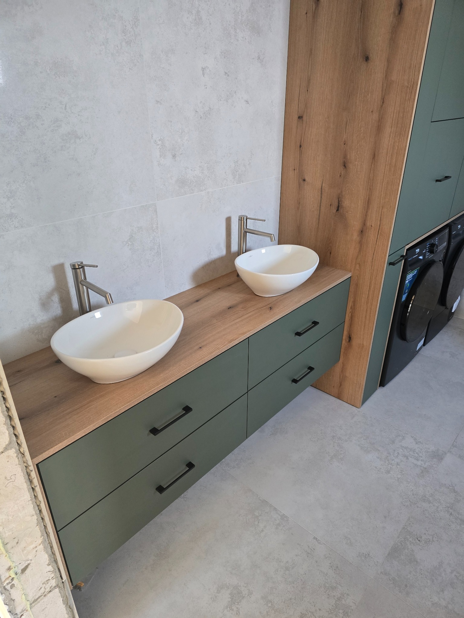 Nowoczesna łazienka z dwiema umywalkami nablatowymi na drewnianym blacie i zielonymi szafkami. Obok zabudowa z pralką i suszarką. Minimalistyczny design, jasne płytki.