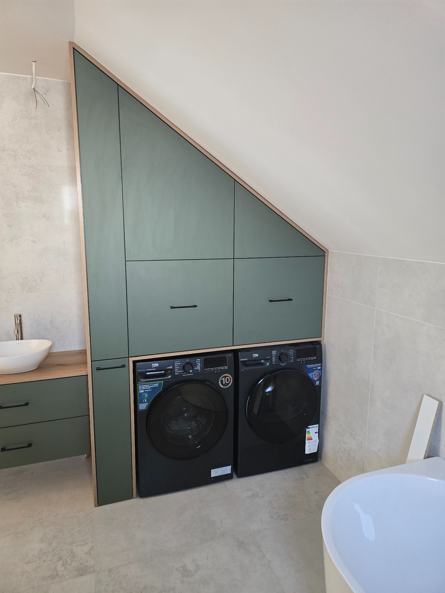 Zielona zabudowa meblowa w łazience na poddaszu, z szafkami i miejscem na pralkę i suszarkę. Umywalka nablatowa i płytki w tle.