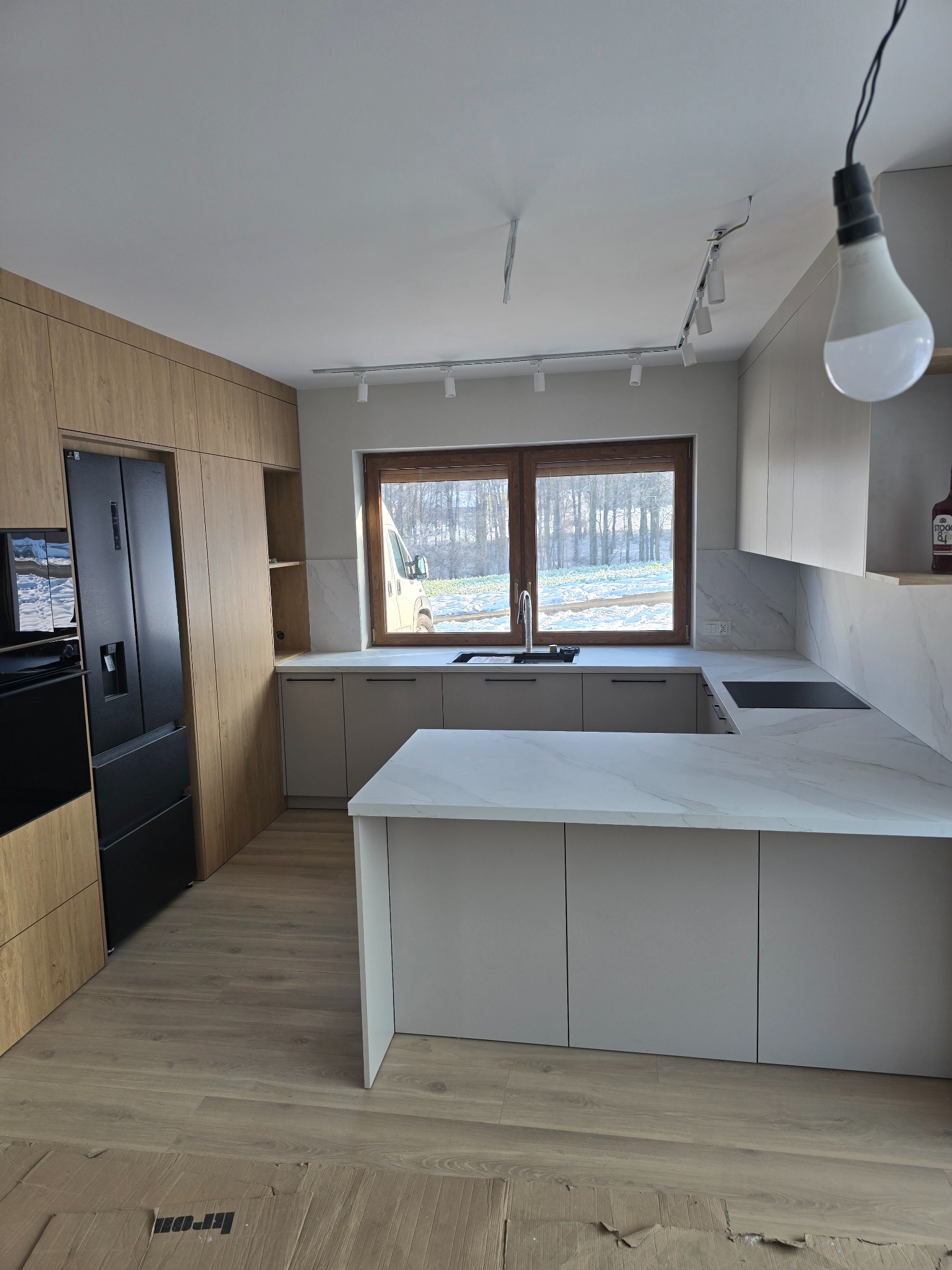 Nowoczesna kuchnia z wyspą, fronty w odcieniach beżu i drewna, marmurowy blat, czarna lodówka, oświetlenie szynowe. Widok na zimowy krajobraz za oknem.