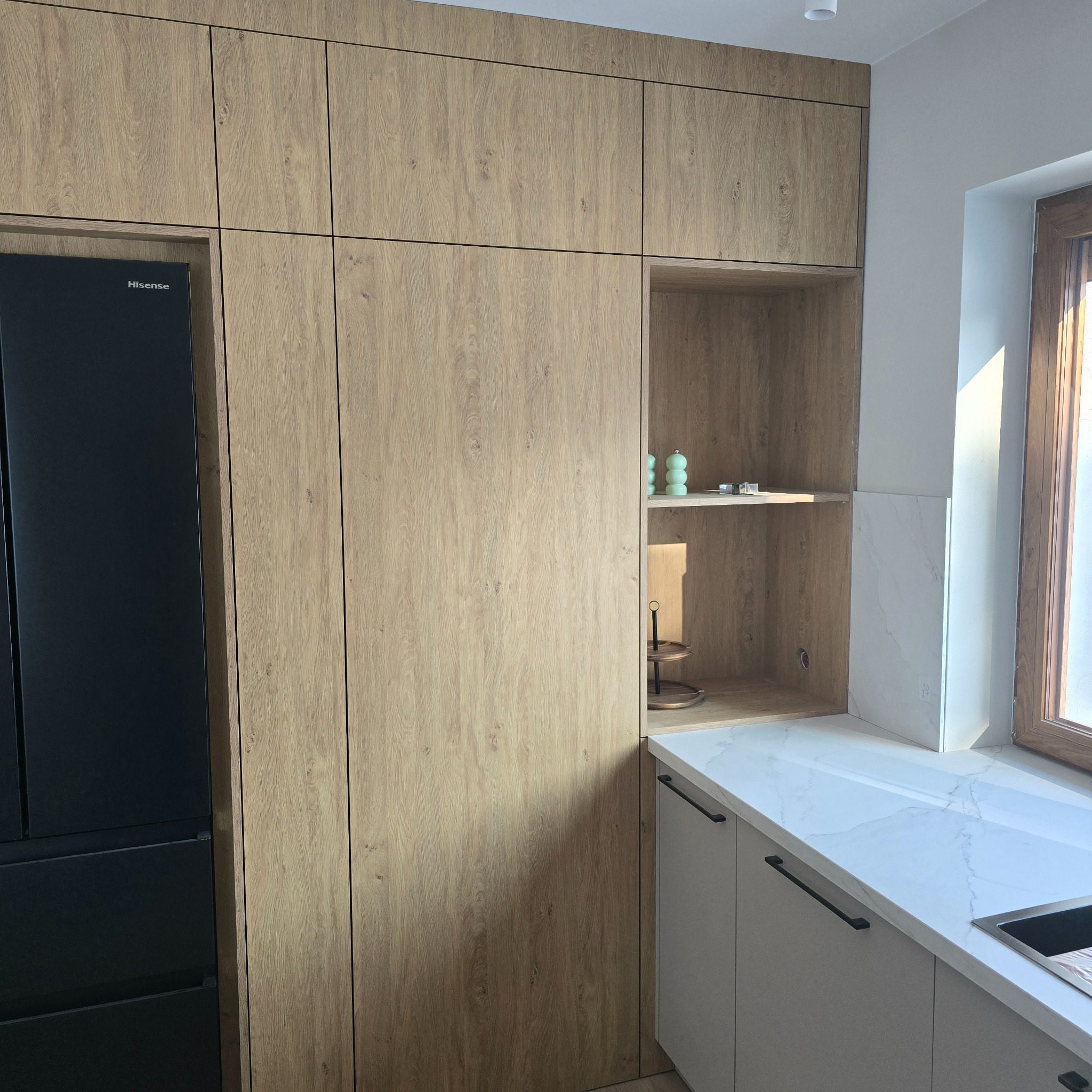 Jasna, drewnopodobna zabudowa kuchenna z czarną lodówką Hisense i otwartą półką z dekoracjami. Biały blat i okno z drewnianą ramą w tle.