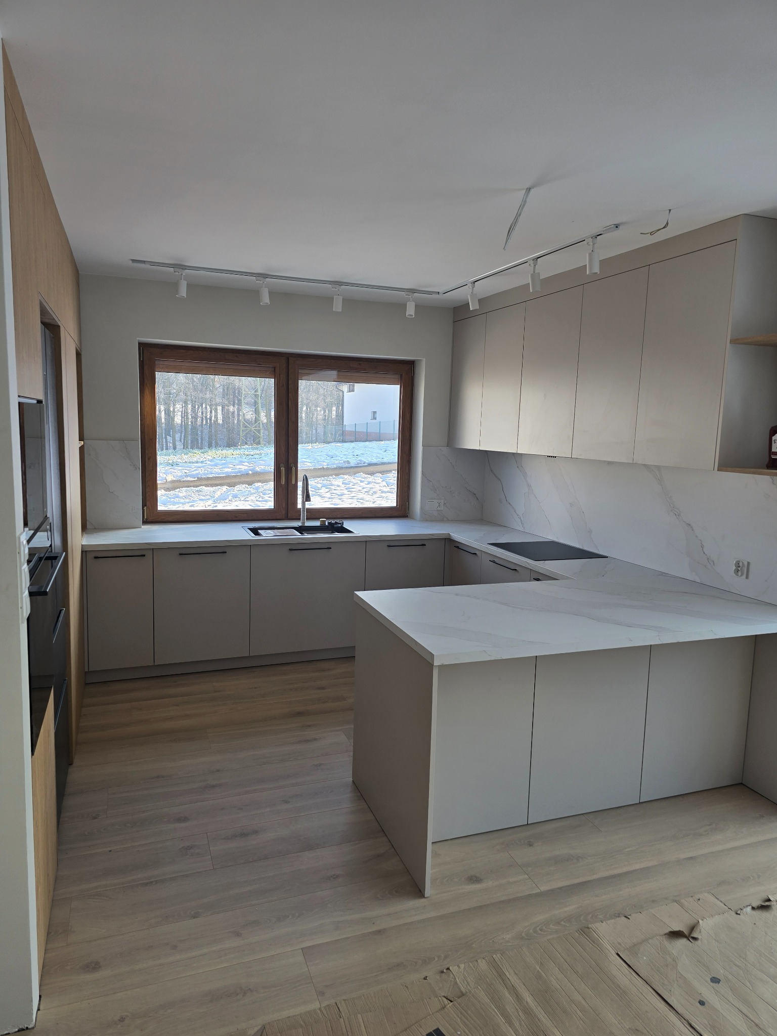 Jasna kuchnia z wyspą i marmurowym blatem, oświetlona reflektorami. Za oknem zimowy krajobraz. Minimalistyczny design z drewnianymi akcentami.