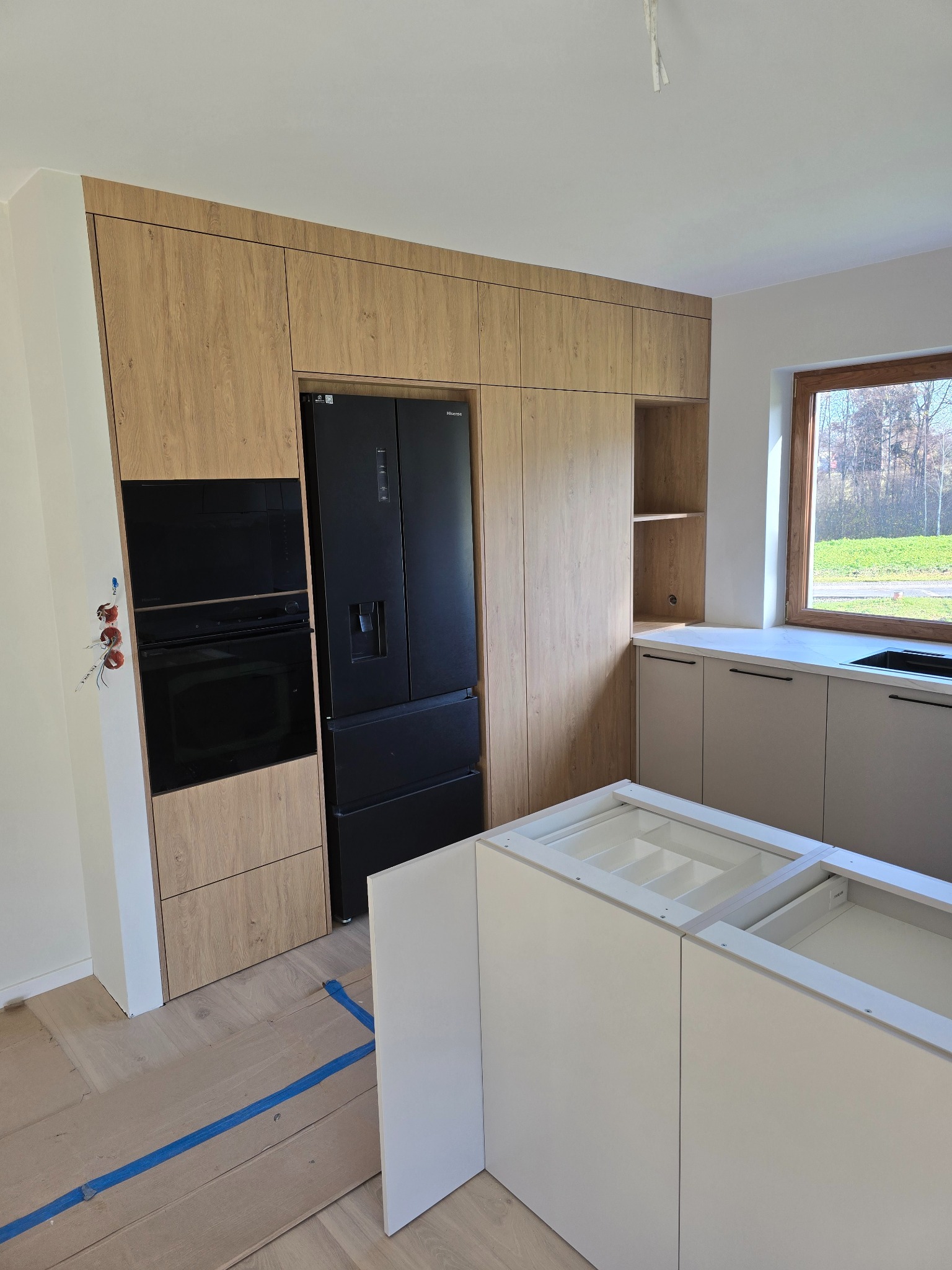 Jasna kuchnia z drewnianymi frontami, czarnym piekarnikiem i lodówką. Częściowo zmontowana wyspa kuchenna na pierwszym planie. Widoczne instalacje elektryczne.