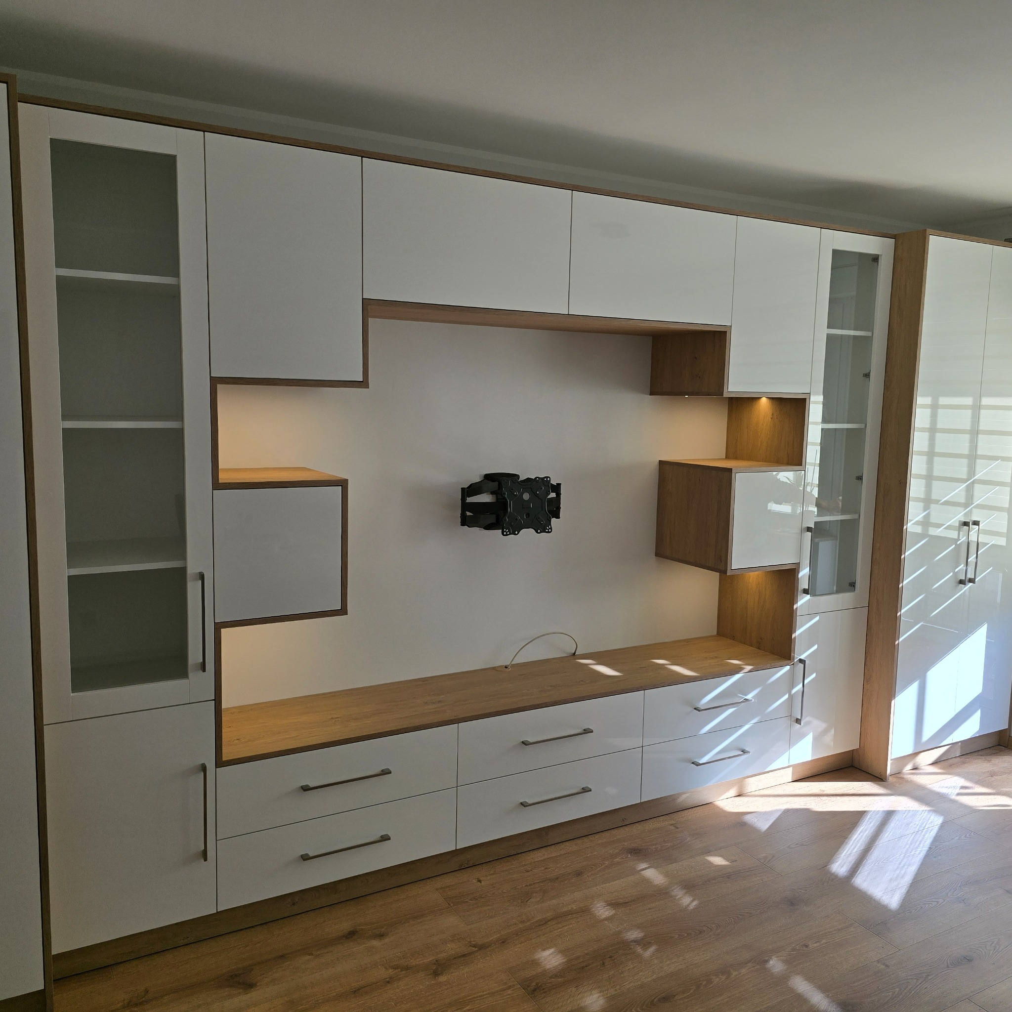 Biała zabudowa meblowa z drewnianymi akcentami i oświetleniem LED, z uchwytem na telewizor na ścianie. Nowoczesny design, widok z perspektywy podłogi.