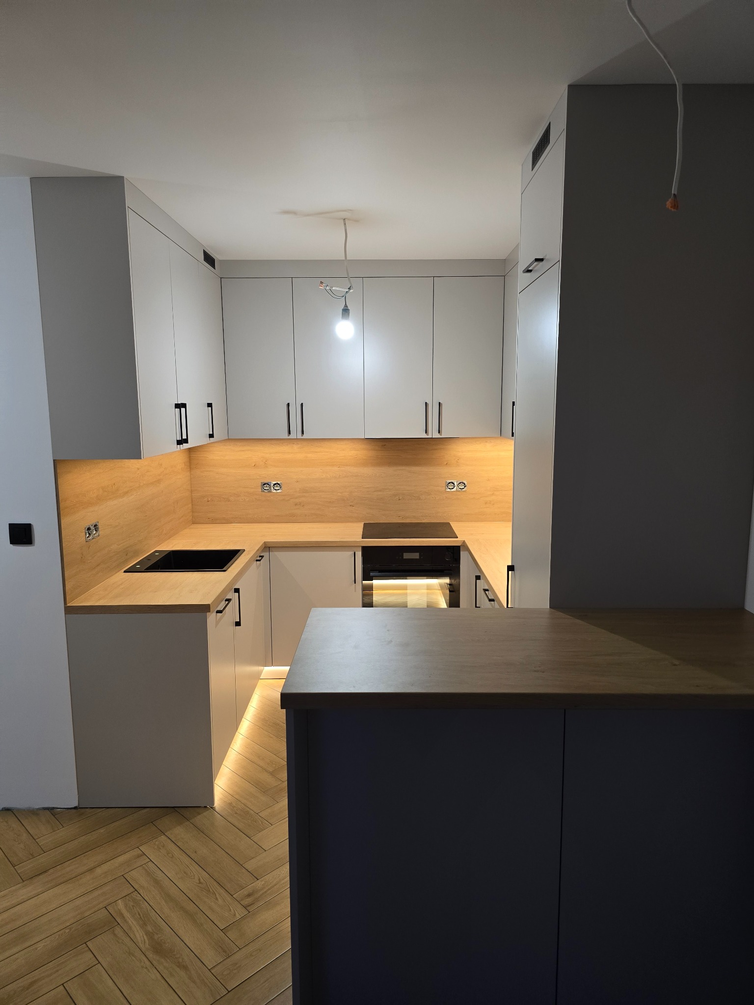 Nowoczesna kuchnia w kształcie litery U z szarymi szafkami, drewnianym blatem i podświetleniem LED. Widoczna płyta indukcyjna i fragment podłogi w jodełkę.