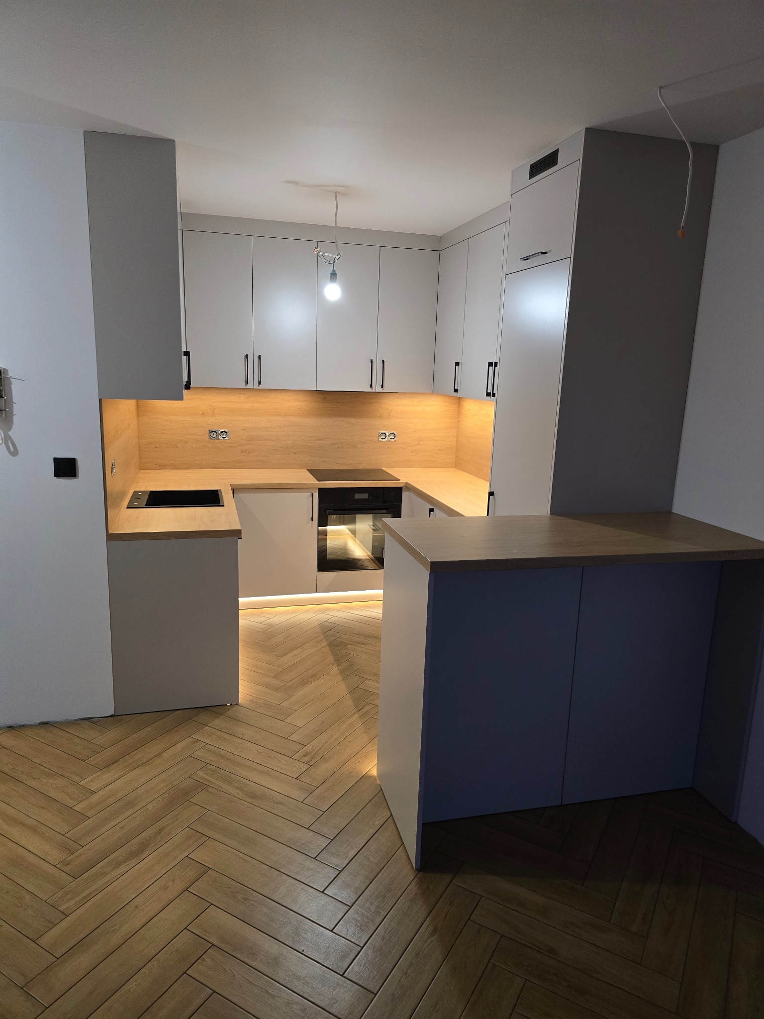 Nowoczesna kuchnia z szarymi szafkami, drewnianym blatem i podświetleniem LED pod szafkami. Parkiet ułożony w jodełkę. Widoczny fragment wyspy kuchennej.