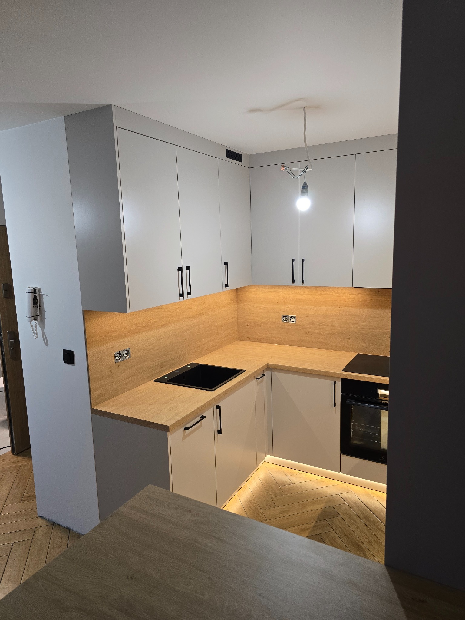 Jasna kuchnia z drewnianym blatem i czarnymi uchwytami. Oświetlenie LED pod szafkami dolnymi. Parkiet w jodełkę. Minimalistyczny design.