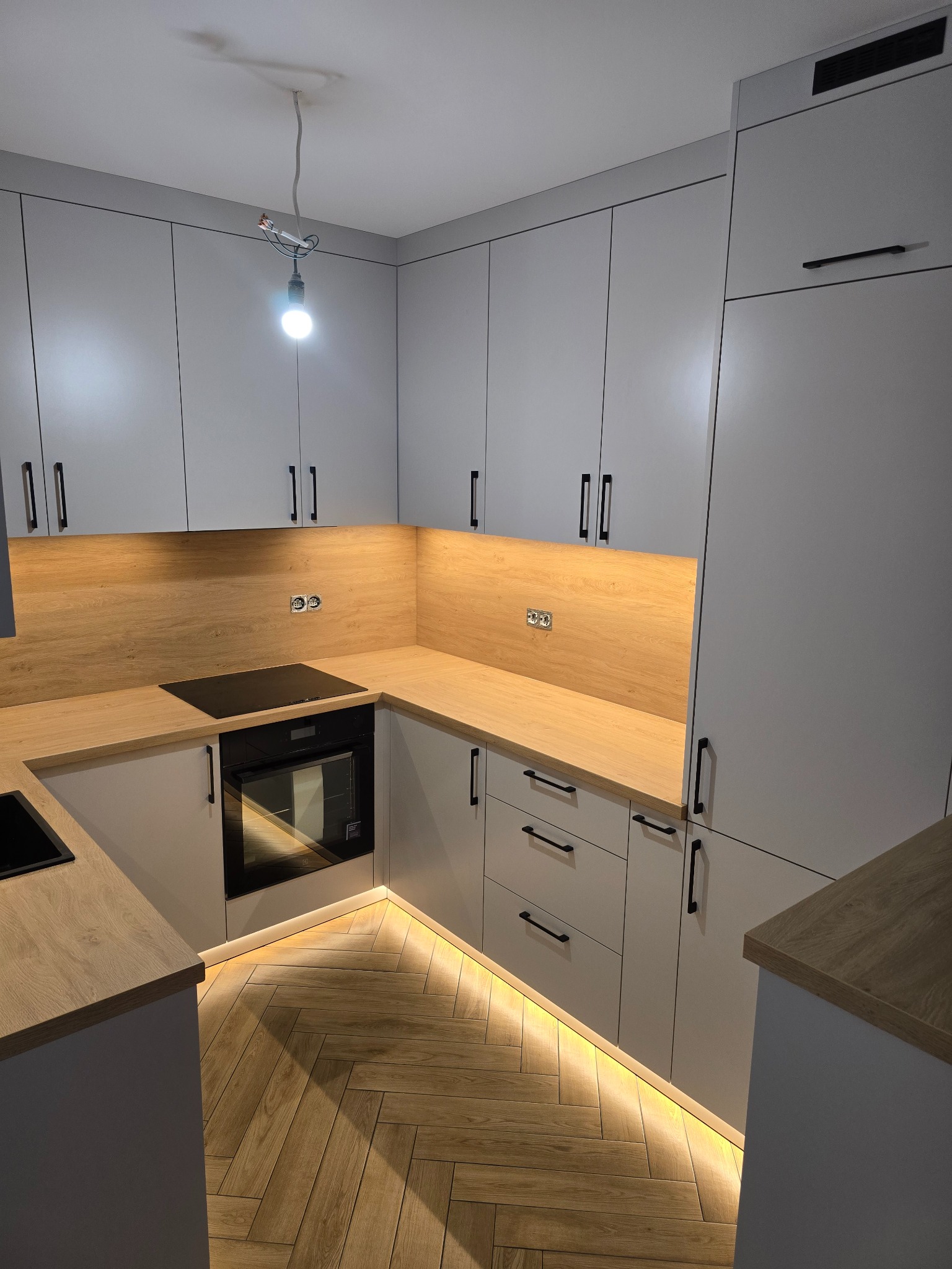 Szara kuchnia z drewnianym blatem i podświetleniem LED przy podłodze. Uwagę przykuwają czarne, minimalistyczne uchwyty szafek i parkiet ułożony w jodełkę.