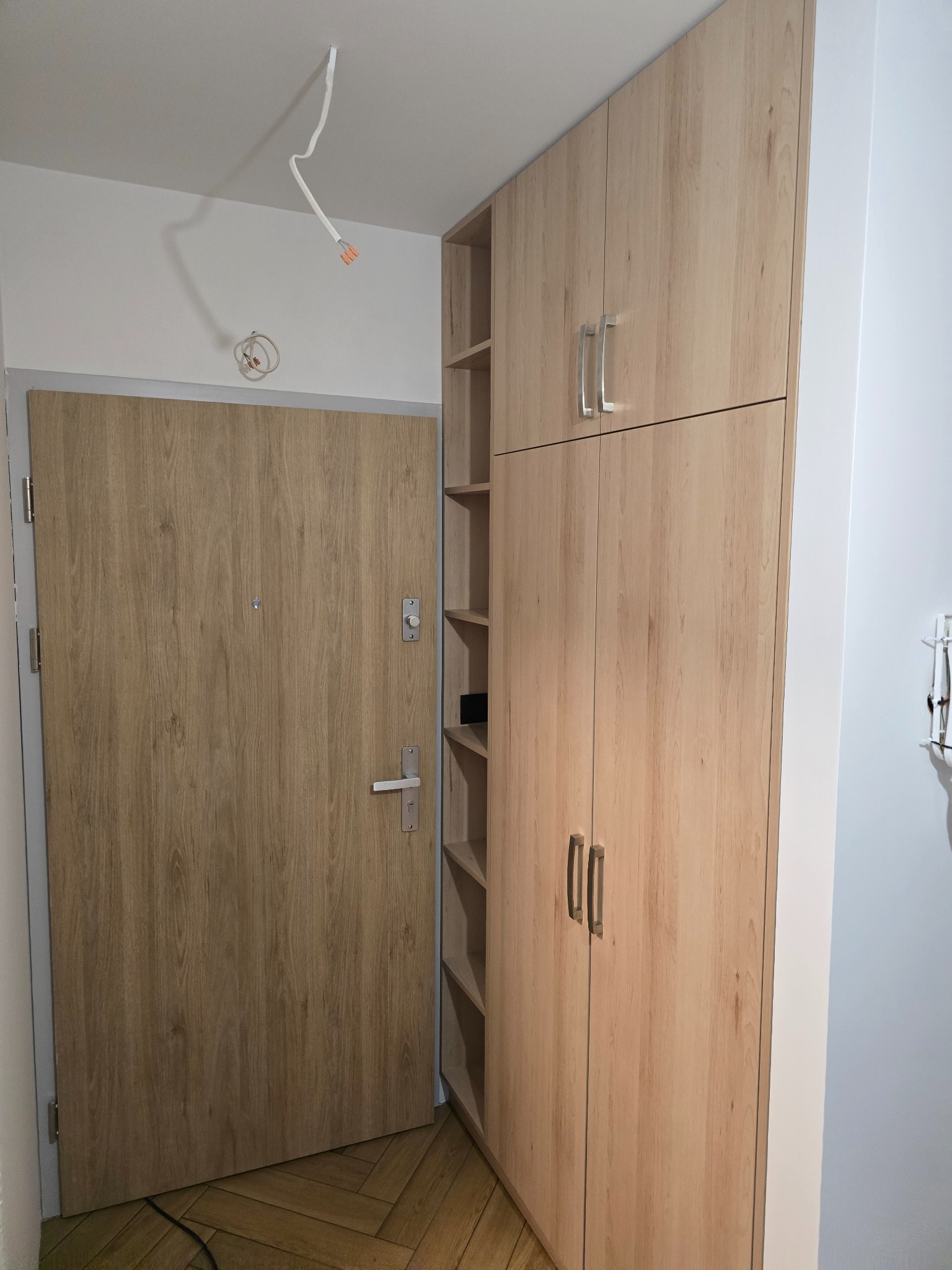 Jasna szafa wnękowa z półkami obok drzwi wejściowych, dopasowana kolorystycznie do drzwi. Minimalistyczny design, widoczne metalowe uchwyty.