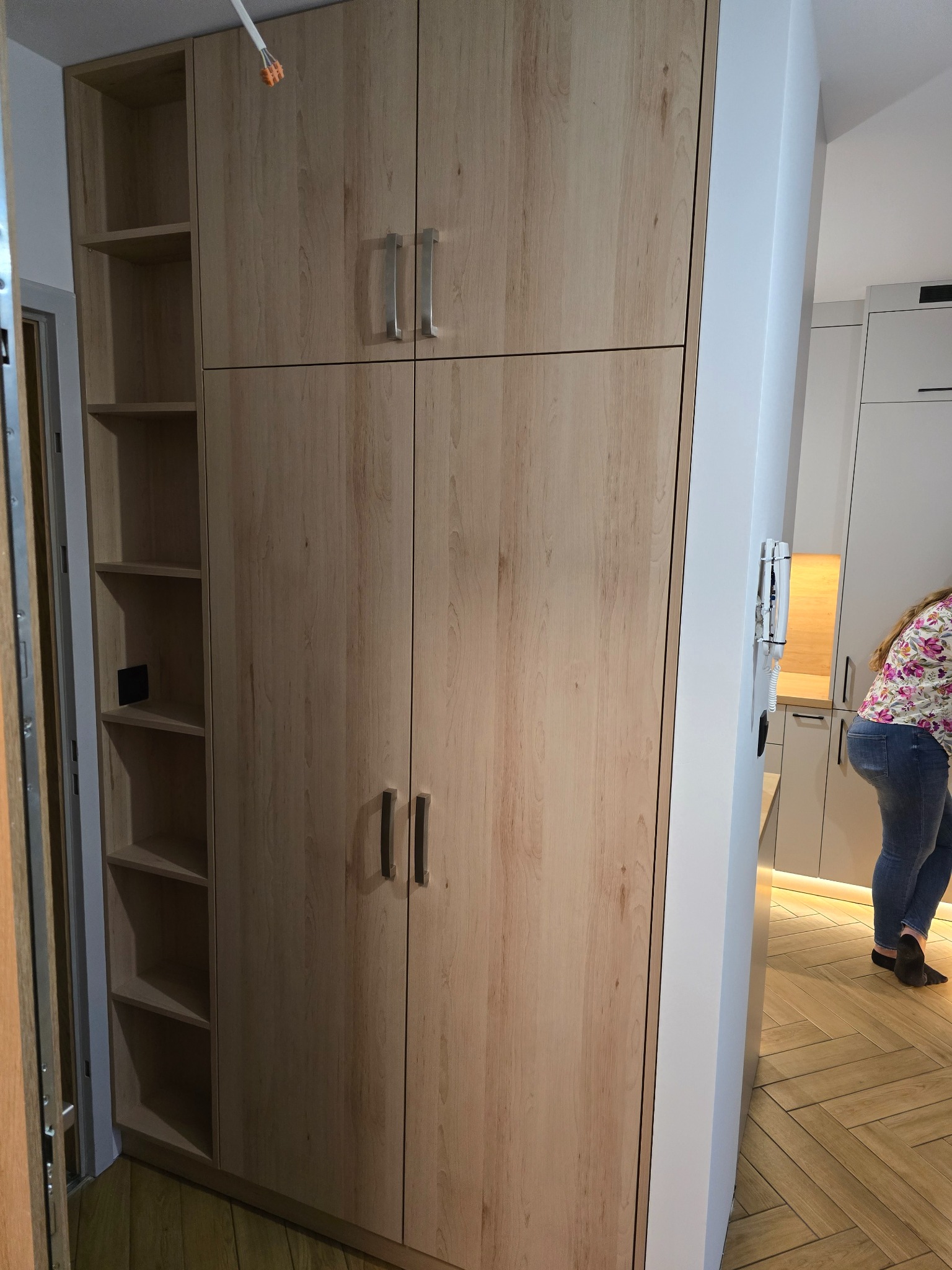 Jasna, drewnopodobna szafa z półkami i drzwiami, dopasowana do wnętrza z parkietem w jodełkę. Minimalistyczne uchwyty i oświetlenie LED w tle.