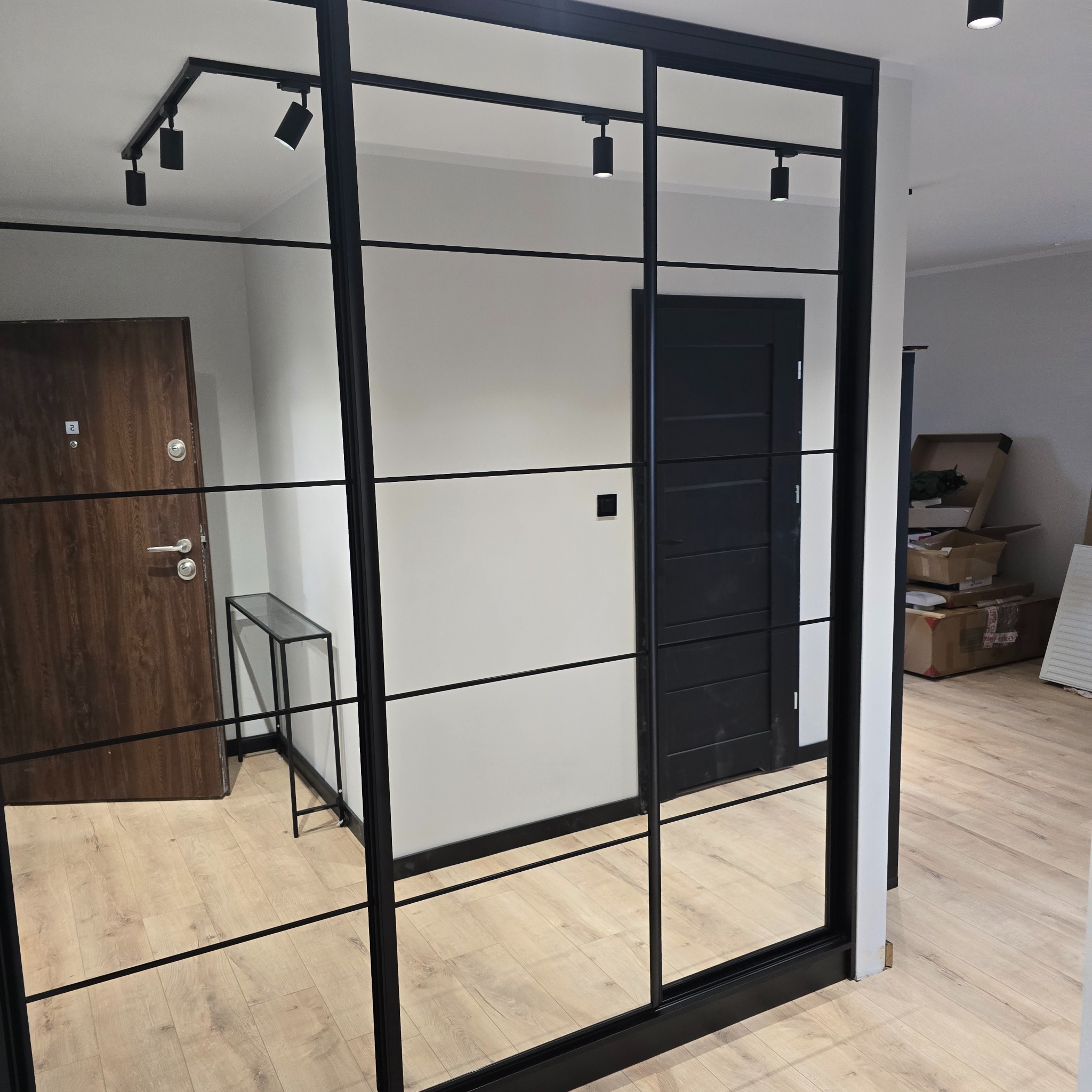 Nowoczesna szafa wnękowa z lustrzanymi drzwiami przesuwnymi i czarnymi profilami, oświetlenie punktowe na suficie, widok na przedpokój z drewnianymi drzwiami.