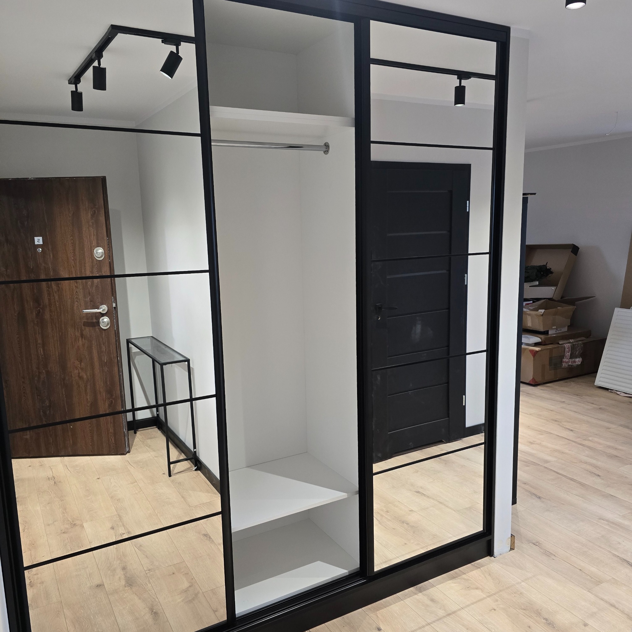 Nowoczesna szafa przesuwna z czarnymi ramami i lustrzanymi drzwiami, widok na wnętrze z półkami i drążkiem, oświetlenie sufitowe, drzwi w tle.