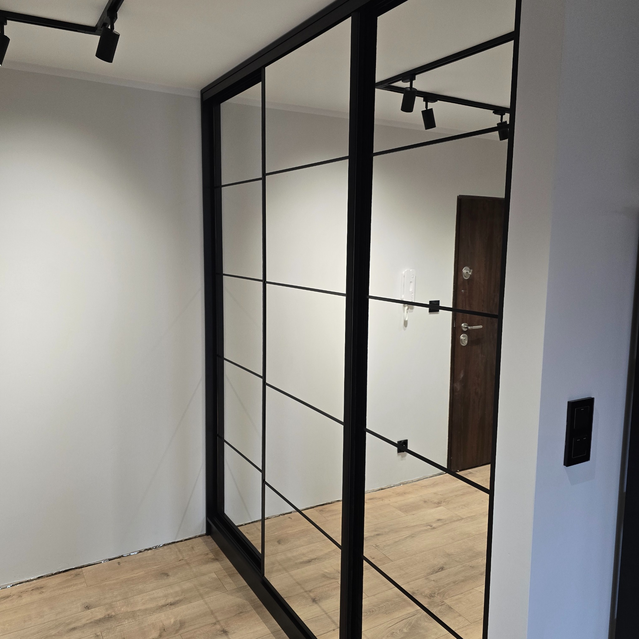 Nowoczesna szafa wnękowa z lustrzanymi drzwiami i czarnymi profilami, odbijająca wnętrze pokoju z drewnianą podłogą i oświetleniem szynowym.