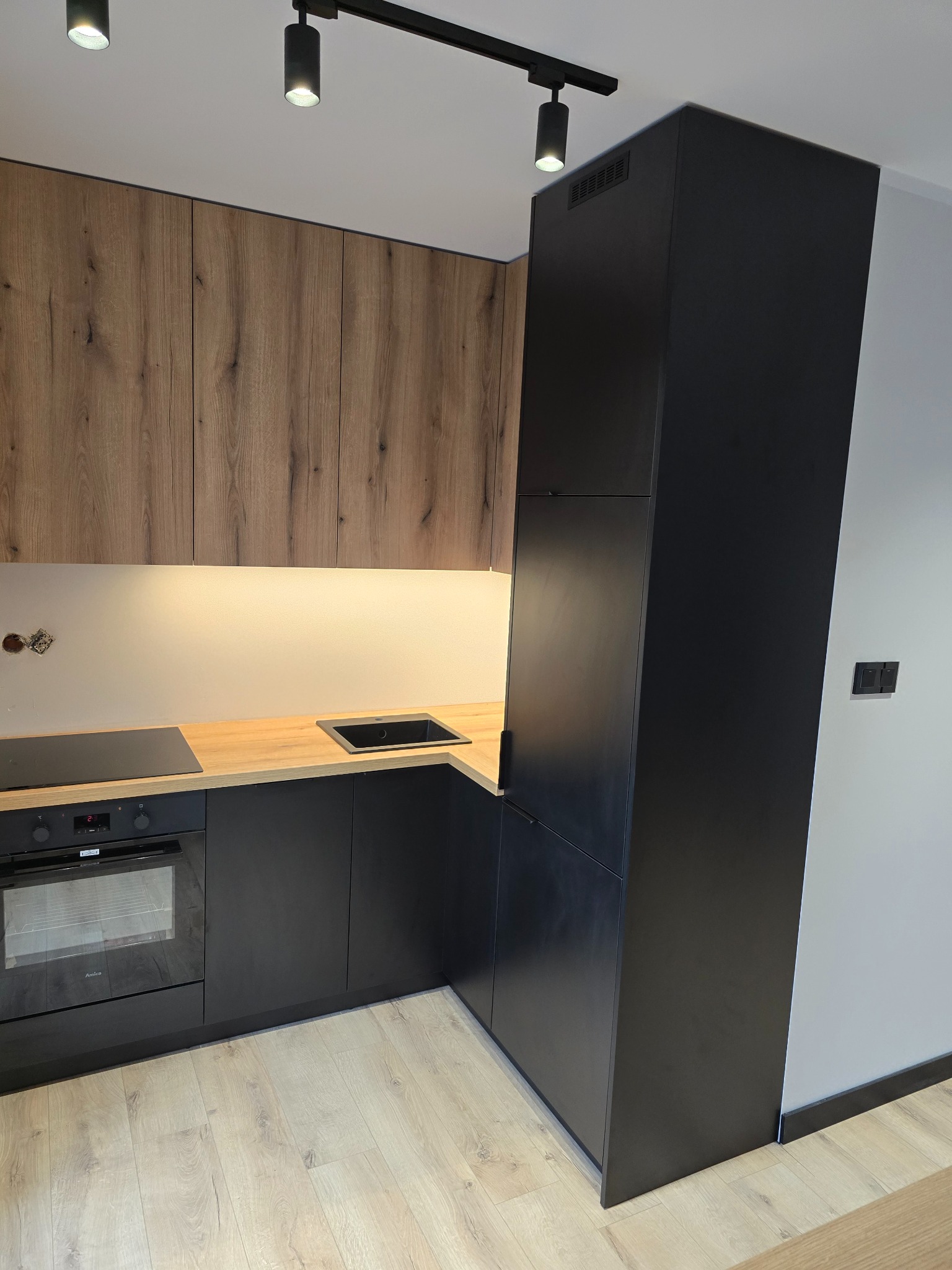 Nowoczesna kuchnia z drewnianymi szafkami górnymi i czarnymi dolnymi, oświetleniem punktowym i drewnianym blatem. Minimalistyczny design z piekarnikiem Amica.