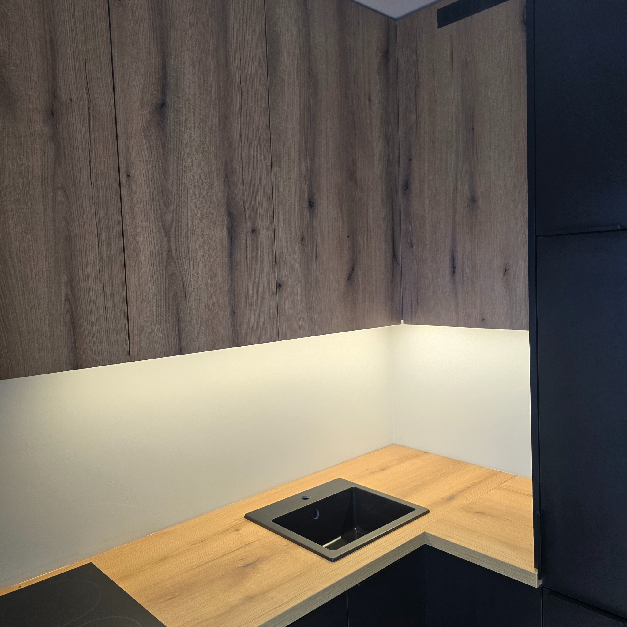 Fragment nowoczesnej kuchni: drewniane szafki wiszące, blat z wpuszczonym czarnym zlewem i oświetlenie LED pod szafkami. Minimalistyczny design.