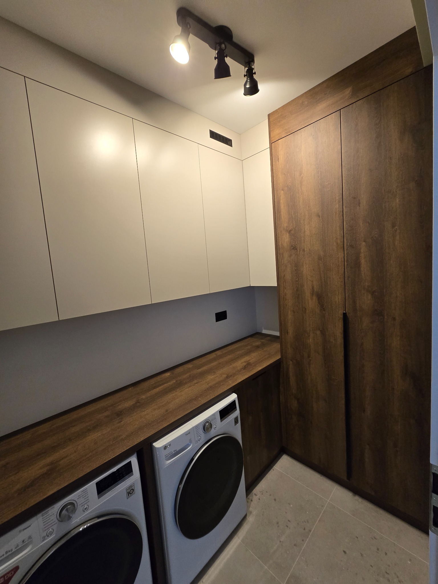 Nowoczesna aranżacja pralni z zabudową meblową: białe szafki wiszące, drewniany blat nad pralkami i wysoka szafa w odcieniu ciemnego drewna. Oświetlenie punktowe.