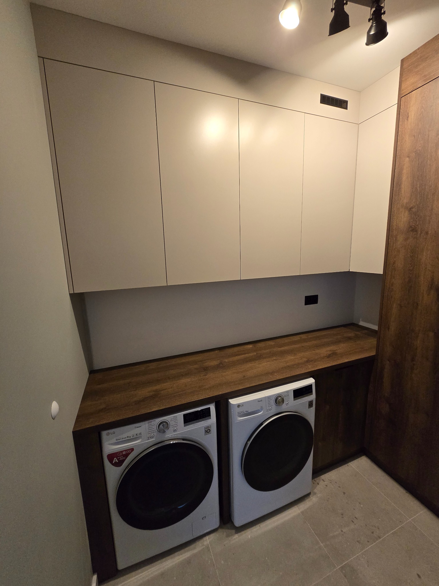 Jasne szafki wiszące nad pralkami LG, blat z ciemnego drewna, oświetlenie punktowe. Minimalistyczny design pralni z elementami drewnianymi.