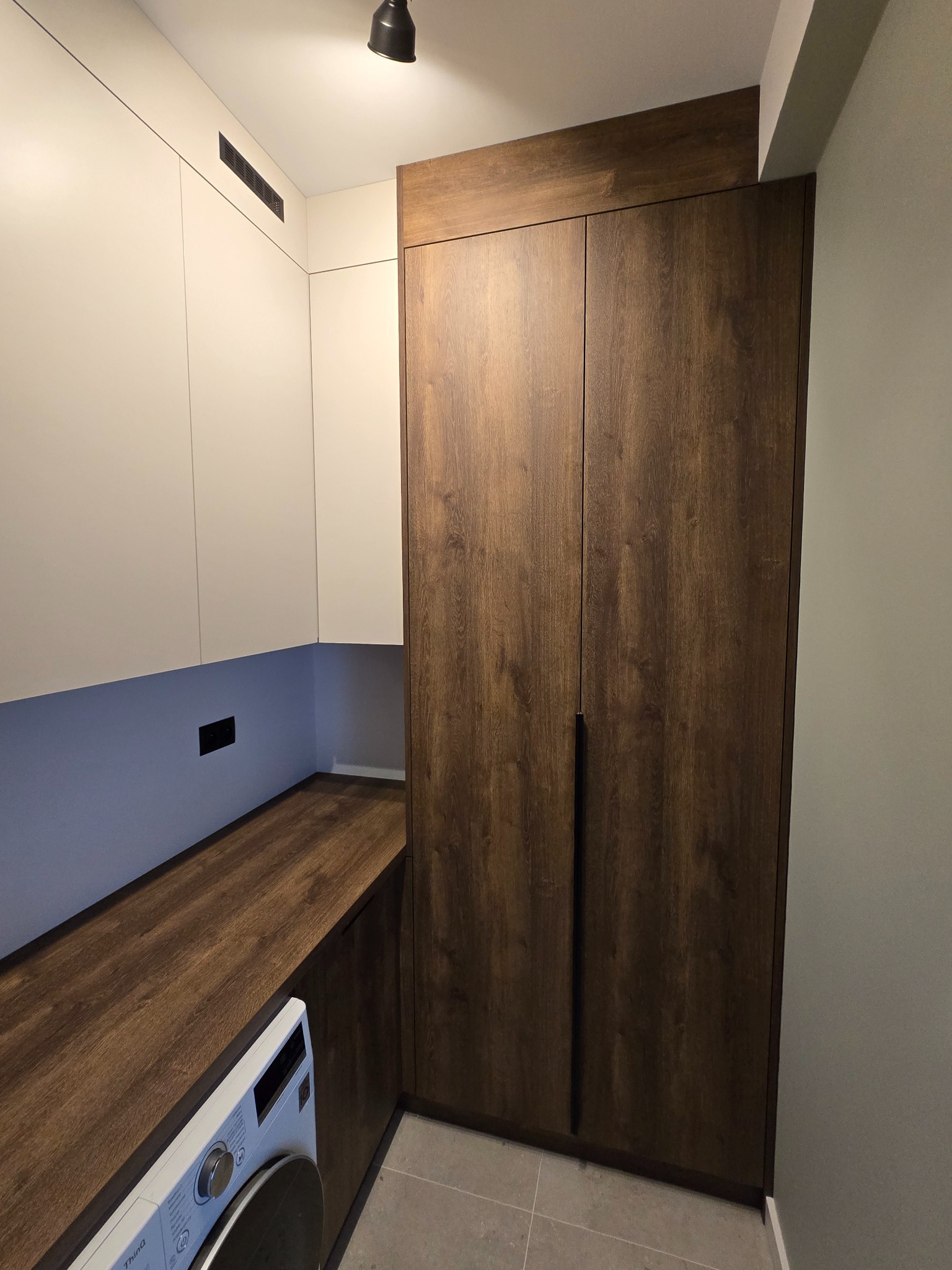 Nowoczesna zabudowa: szafa z ciemnego drewna i białe szafki wiszące z wentylacją, uzupełnione blatem z tego samego drewna. Pralka częściowo widoczna.