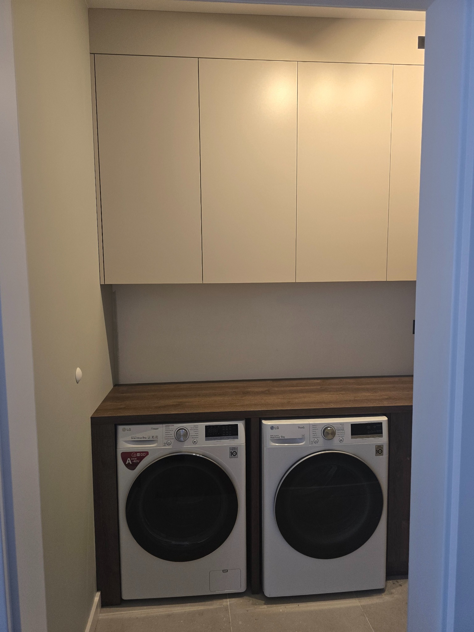 Białe szafki nad pralkami LG w zabudowie z blatem z ciemnego drewna. Minimalistyczny design, nowoczesne rozwiązanie do pralni lub łazienki.