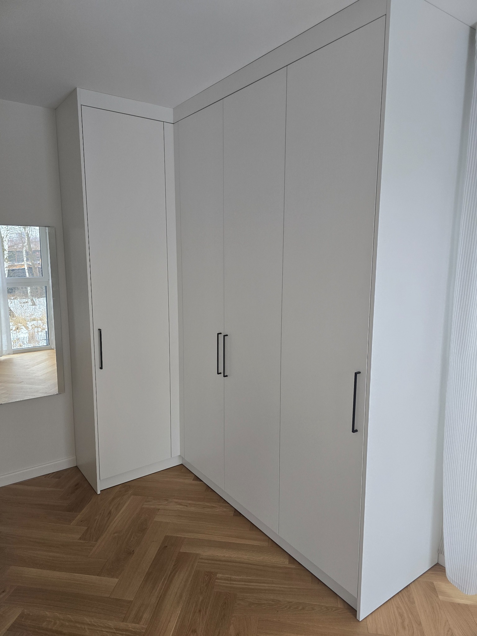 Biała, narożna szafa wnękowa z minimalistycznymi, czarnymi uchwytami, na parkiecie ułożonym w jodełkę. W tle widoczne okno i lustro.