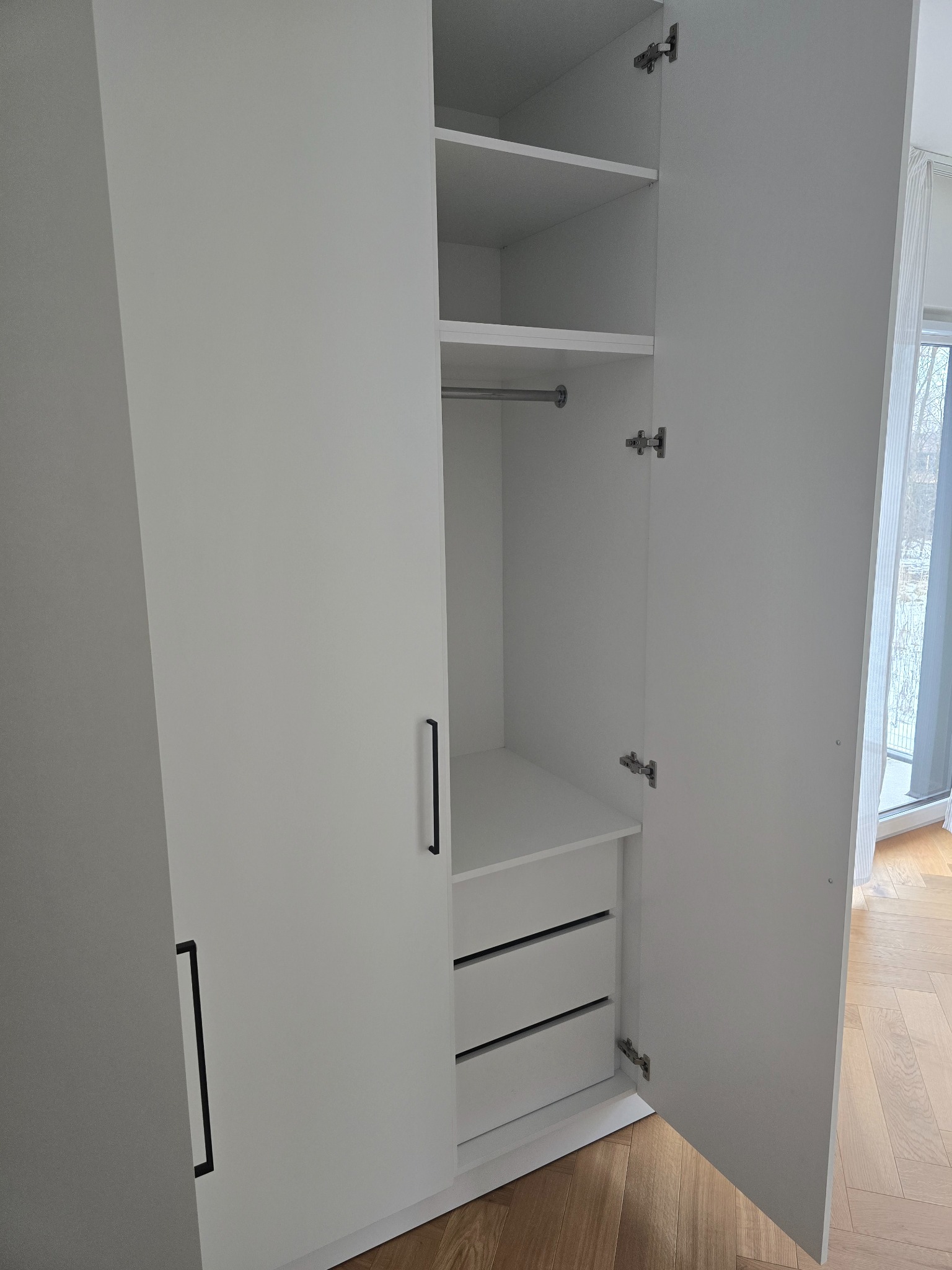Biała szafa z otwartymi drzwiami, widoczne półki, drążek na ubrania i szuflady. Minimalistyczny design, jasne wnętrze, parkiet na podłodze.