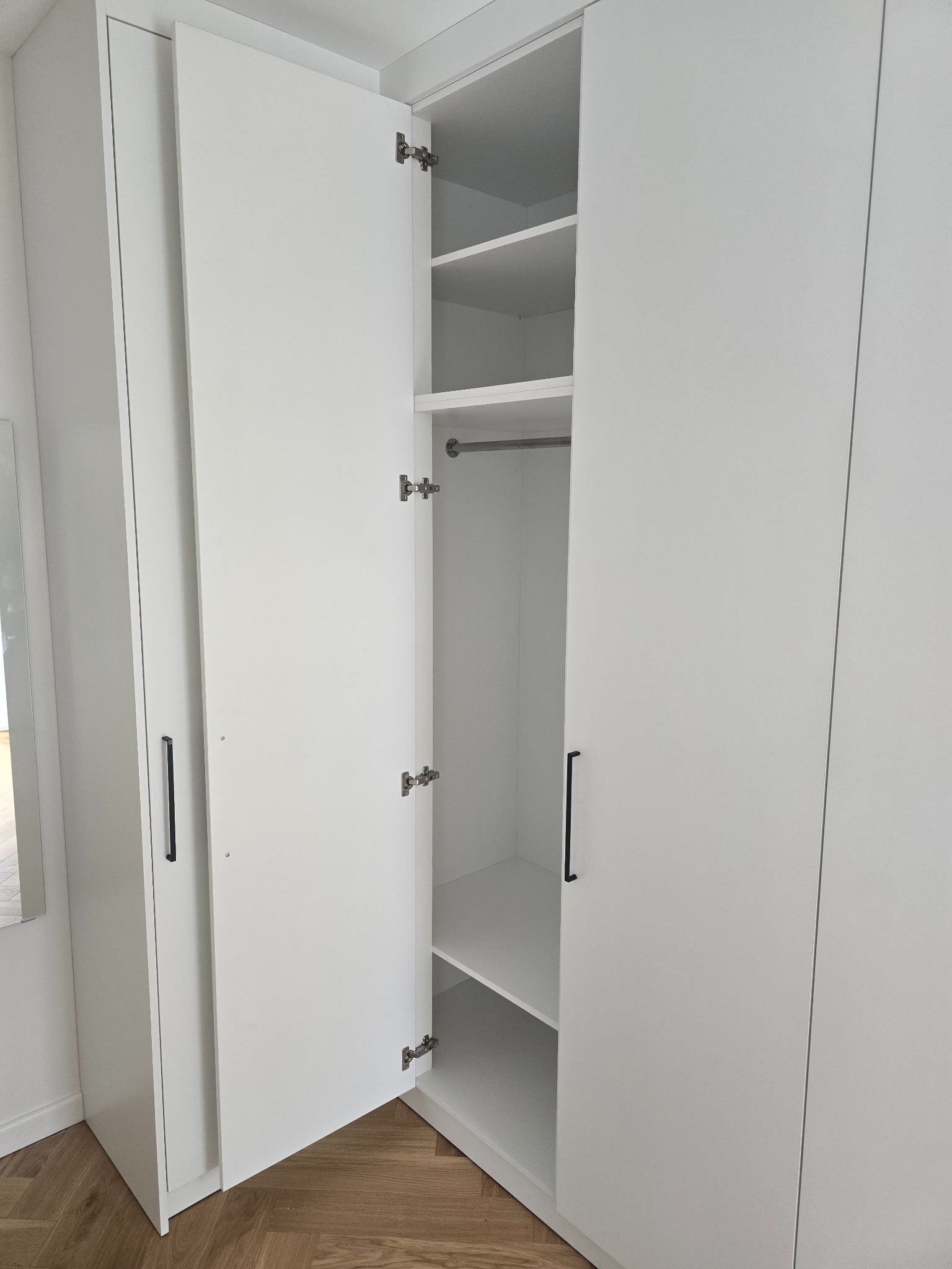 Biała szafa wnękowa z otwartymi drzwiami, widoczne półki i drążek na ubrania. Minimalistyczny design, czarne uchwyty, parkiet na podłodze.