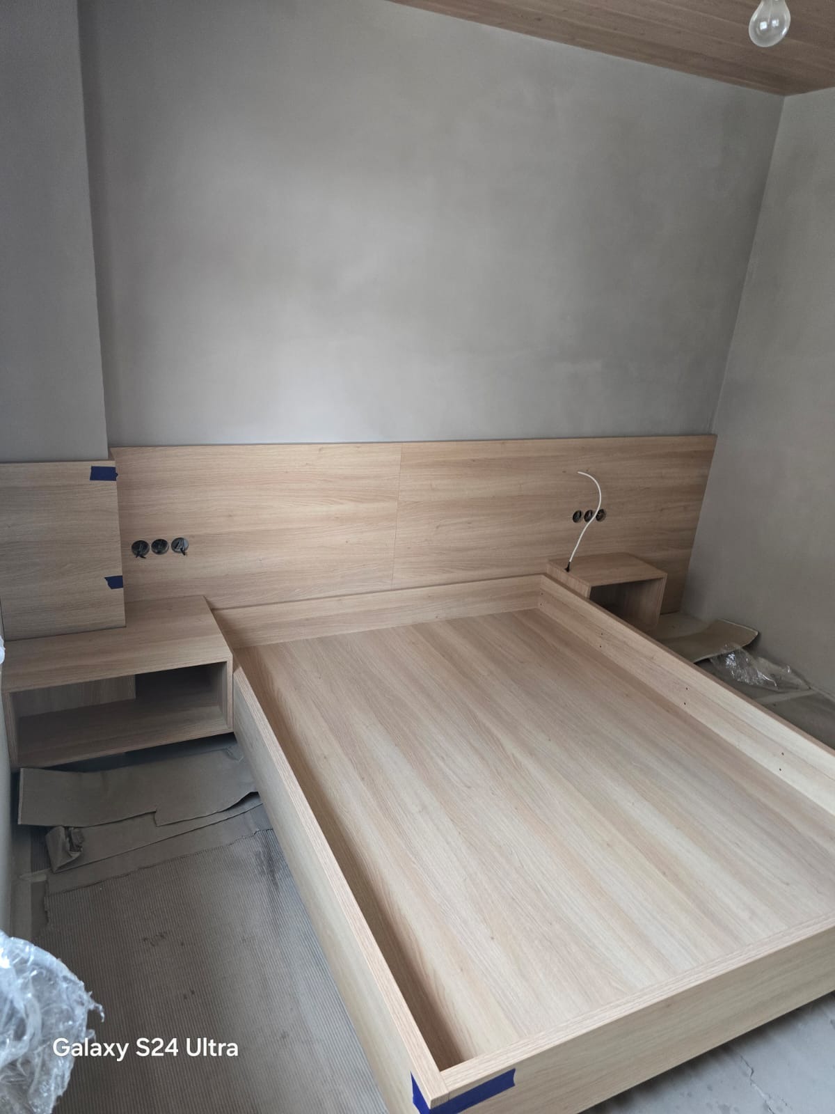 Drewniane łóżko z szafkami nocnymi w surowym wnętrzu. Widoczne gniazdka elektryczne i fragmenty folii ochronnej. Jasne drewno, minimalistyczny design.